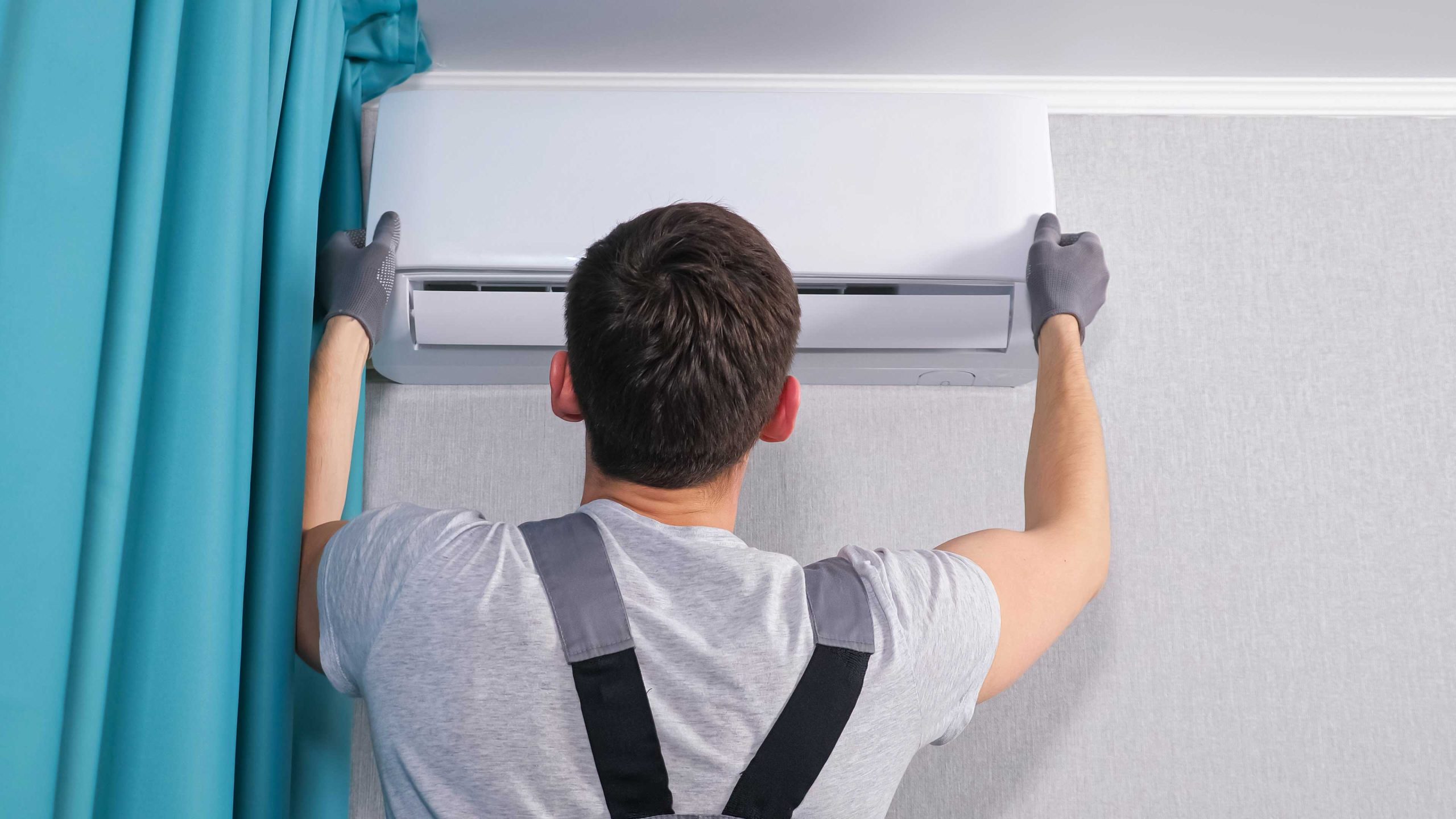 electrician-workwear-opens-air-conditioner-unit-lid-takes-out-screws-with-screwdriver-spacious-apartment-room-close-backside-view